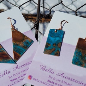 Peut inclure: Deux paires de boucles d'oreilles sur des cartes blanches avec le texte "Bella Accessories by Julie Misasi Made in Canada". Les boucles d'oreilles sont en cuivre avec un motif marbré bleu et brun. Une paire est rectangulaire et l'autre est triangulaire.