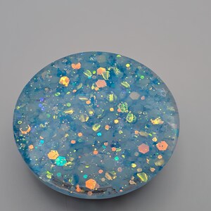 May include: A circular resin disc featuring a translucent light blue base embedded with a variety of iridescent, holographic, and metallic glitter particles in shades of blue, orange, and silver that catch the light to create a shimmering effect.