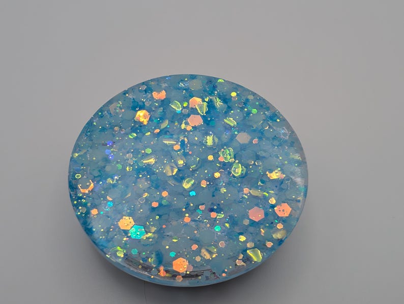 May include: A circular resin disc featuring a translucent light blue base embedded with a variety of iridescent, holographic, and metallic glitter particles in shades of blue, orange, and silver that catch the light to create a shimmering effect.