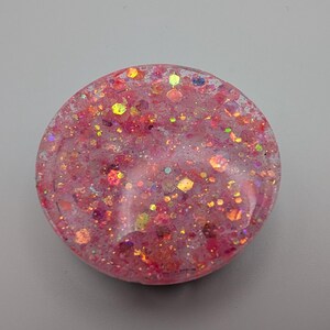 May include: A circular resin piece featuring a translucent pink base embedded with a dense mixture of multi-colored glitter, including hexagonal and fine particles in shades of gold, orange, and magenta that catch the light across the surface.