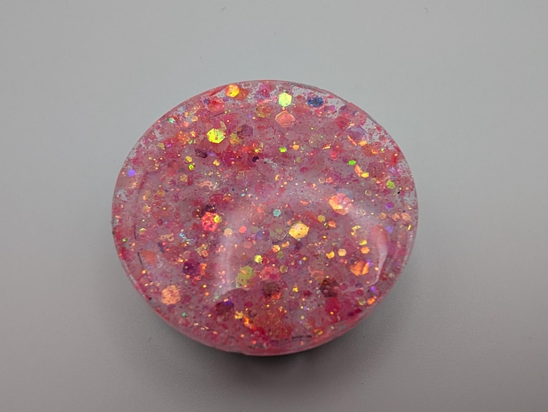 May include: A circular resin piece featuring a translucent pink base embedded with a dense mixture of multi-colored glitter, including hexagonal and fine particles in shades of gold, orange, and magenta that catch the light across the surface.