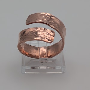 May include: A copper-coloured, hammered metal bracelet with an open, wrap-around design. The bracelet has a textured surface and is displayed on a clear, rectangular stand. The metal has a warm, reddish-brown hue.