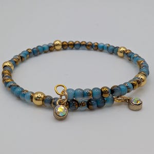 Puede incluir: Una pulsera envolvente con cuentas azules y doradas y detalles dorados. La pulsera tiene un cierre dorado y un pequeño dije con una piedra iridiscente. La pulsera está diseñada para envolverse alrededor de la muñeca.