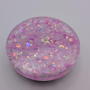 May include: A circular, pink resin object filled with pink and iridescent glitter. The glitter reflects light, creating a sparkling effect. The object is a solid, circular shape. The glitter is of various sizes.