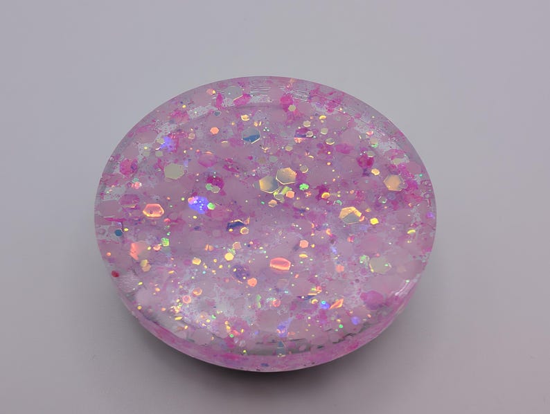 May include: A circular resin coaster featuring a translucent light pink base embedded with a variety of iridescent, holographic, and magenta glitter flakes. The surface is smooth and glossy, showcasing a dense distribution of reflective hexagonal and circular shapes.