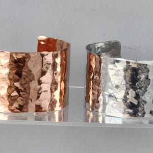 Pode incluir: Duas pulseiras de metal martelado, uma de cobre e outra de prata. Ambas as pulseiras são largas e têm uma superfície texturizada.