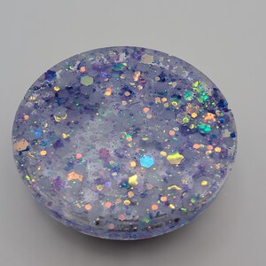 May include: A circular resin coaster featuring a translucent light purple base embedded with a variety of multi-colored hexagonal and fine glitter particles in shades of blue, pink, and iridescent silver that catch the light across the surface.