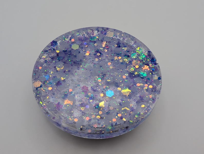 May include: A circular resin coaster featuring a translucent light purple base embedded with a variety of multi-colored hexagonal and fine glitter particles in shades of blue, pink, and iridescent silver that catch the light across the surface.