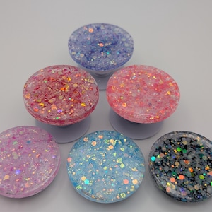 May include: A collection of five circular phone grips arranged in a pyramid formation. Each grip features a unique resin surface embedded with various sizes of colorful, reflective glitter in shades of purple, pink, light blue, and dark gray.