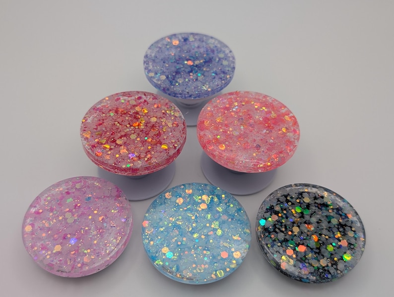 May include: A collection of five circular phone grips arranged in a pyramid formation. Each grip features a unique resin surface embedded with various sizes of colorful, reflective glitter in shades of purple, pink, light blue, and dark gray.