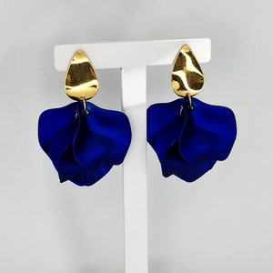 May include: A pair of blue flower-shaped earrings with gold accents. The earrings are hanging from a white display stand.