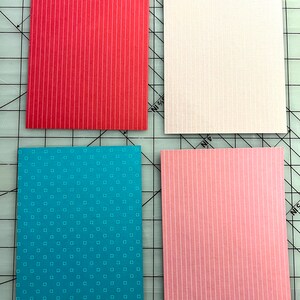 May include: Four rectangular patterned paper sheets. One is red with vertical white stripes, another is white with a subtle grid pattern, a third is teal with small white squares, and the last is pink with vertical white stripes.