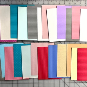 May include: A collection of colorful cardstock paper in various sizes and patterns. The assortment includes solid colors, stripes, dots, and grid designs. The papers are arranged on a gray cutting mat with inch measurements.