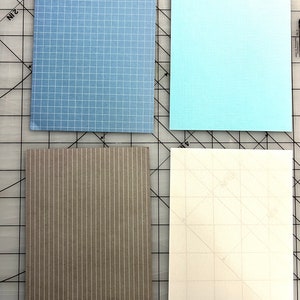 May include: Four rectangular paper sheets in shades of blue, gray, and white. The top left sheet is light blue with a grid pattern. The top right sheet is a solid light blue. The bottom left sheet is gray with vertical stripes. The bottom right sheet is white with a faint grid.