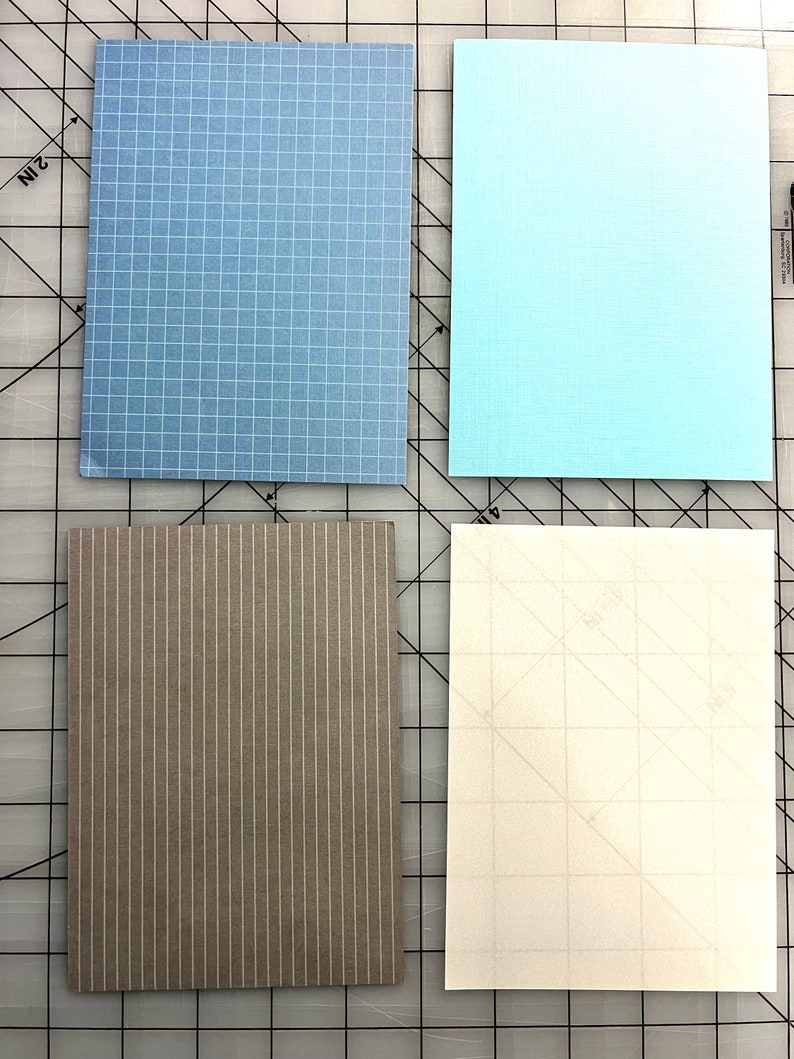 May include: Four rectangular paper sheets in shades of blue, gray, and white. The top left sheet is light blue with a grid pattern. The top right sheet is a solid light blue. The bottom left sheet is gray with vertical stripes. The bottom right sheet is white with a faint grid.