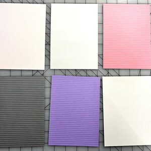 May include: Six rectangular cards in various colors: white, pink, gray, purple, and two more white cards. Each card has a textured surface with subtle horizontal lines. The cards are arranged on a gray cutting mat.