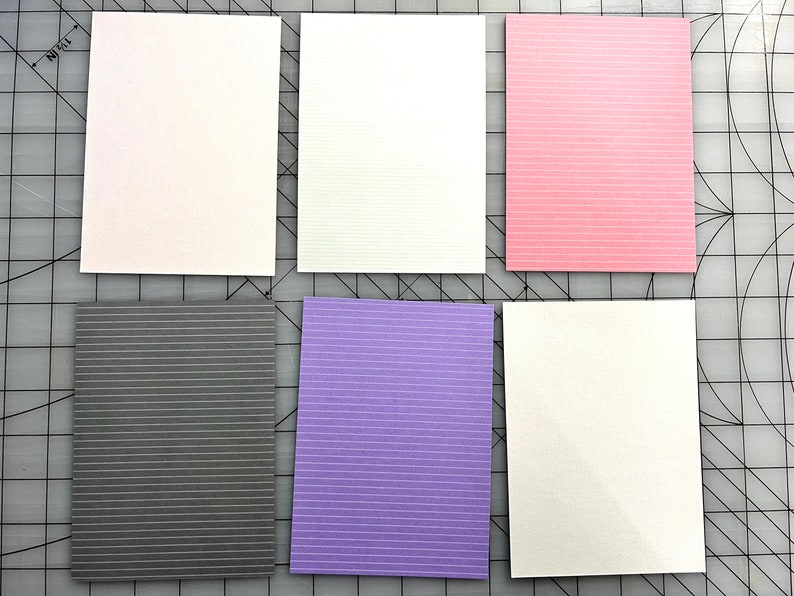 May include: Six rectangular cards in various colors: white, pink, gray, purple, and two more white cards. Each card has a textured surface with subtle horizontal lines. The cards are arranged on a gray cutting mat.