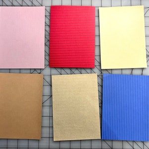 May include: Six rectangular cards in various colors: light pink, red, yellow, brown, beige, and blue. The red and blue cards have a striped texture, while the others have a smooth texture. The cards are arranged on a gray surface.