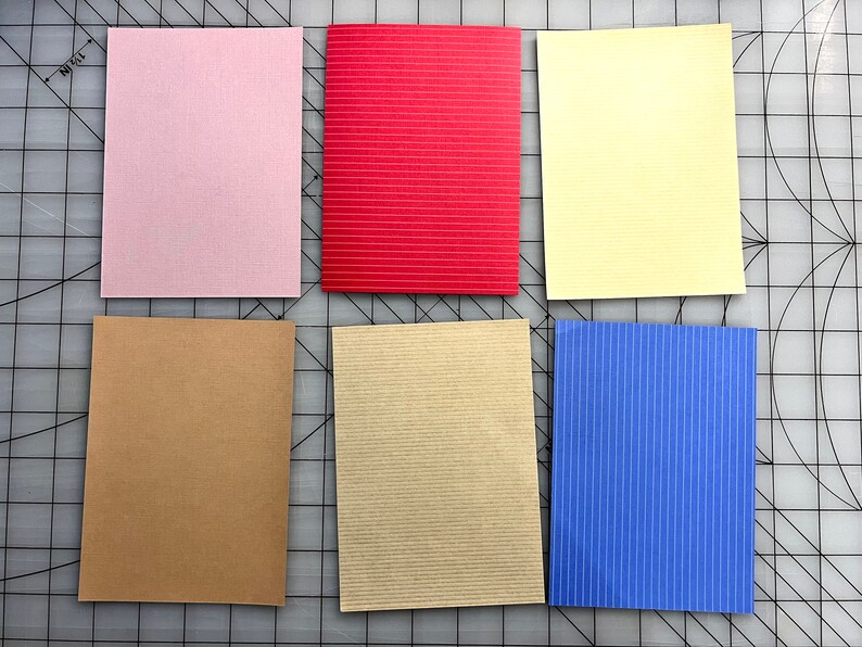 May include: Six rectangular cards in various colors: light pink, red, yellow, brown, beige, and blue. The red and blue cards have a striped texture, while the others have a smooth texture. The cards are arranged on a gray surface.