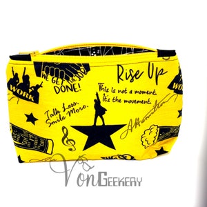 Puede incluir: Bolsa amarilla con cremallera con un estampado en blanco y negro que incluye el texto "Rise Up", "This is not a moment. It's the movement." y "Talk Less. Smile More." La bolsa también presenta una silueta negra de una persona de pie sobre una estrella negra.