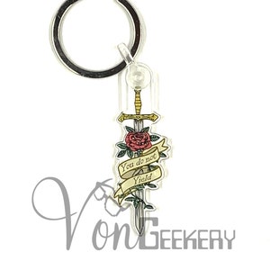 May include: A clear acrylic keychain with a silver metal ring. The keychain features a sword design with a red rose and a banner that reads "You do not yield". The keychain has a gold and silver color scheme.