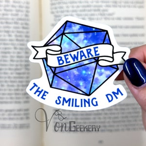 Könnte beinhalten: Ein weißer Aufkleber mit einem blauen und violetten D20-Würfel im Galaxie-Design mit dem Text "Beware the Smiling DM" auf einem weißen Banner.