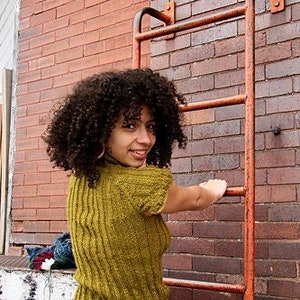 May include: A woman with curly brown hair is wearing a mustard yellow sweater with a ribbed texture. She is standing in front of a brick wall with a metal ladder.