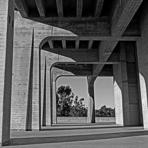 May include: Black and white photograph of a concrete structure, likely a bridge or underpass. The image captures the geometric shapes and shadows created by the architectural design. The composition features repeating pillars and archways, with a view of trees in the background.