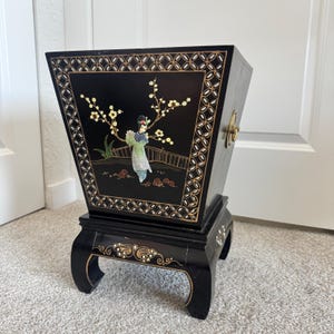 May include: A black tapered wastebasket featuring a hand-painted scene of a figure in traditional attire standing beneath a flowering tree. The piece is accented with gold geometric borders and rests on a four-legged base with gold floral details.