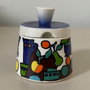 May include: A white ceramic sugar bowl with a blue lid and a colorful abstract design of fruit and geometric shapes. The design includes oranges, pears, and a blue vase.