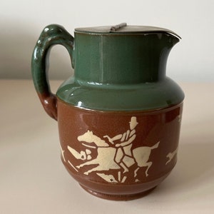 Puede incluir: Una jarra de cerámica marrón y verde con una tapa de peltre. La jarra presenta una silueta blanca de una persona montando a caballo con otros caballos y perros en el fondo.