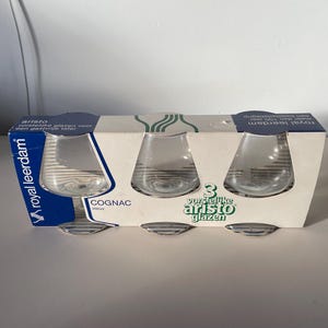 Può includere: Un set di tre bicchieri da cognac in vetro trasparente con un motivo a righe. I bicchieri sono confezionati in una scatola bianca con testo blu e verde che recita "3 vorstelike aristo glazen" e "Royal Leerdam".