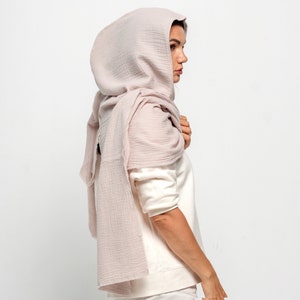 May include: A light mauve-colored scarf and hood combination. The scarf is draped over the shoulders and head, creating a soft, flowing silhouette. The fabric appears to be a lightweight, textured material. The overall look is simple and elegant.