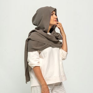 May include: A gray hooded scarf with a waffle texture. The scarf is worn around the neck and head of a person.