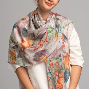 May include: A beige silk scarf with a floral print featuring large orange flowers, green leaves, and blue accents. The scarf is draped around a person's neck.