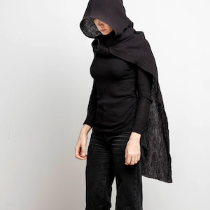 May include: A person wearing a black hooded cloak with a long, flowing design. The hood is pulled up over their head, and they are looking down.