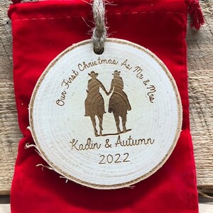 May include: A round wooden ornament with the text "Our First Christmas As Mr & Mrs Kadin & Autumn 2022" and a silhouette of a couple in cowboy hats.