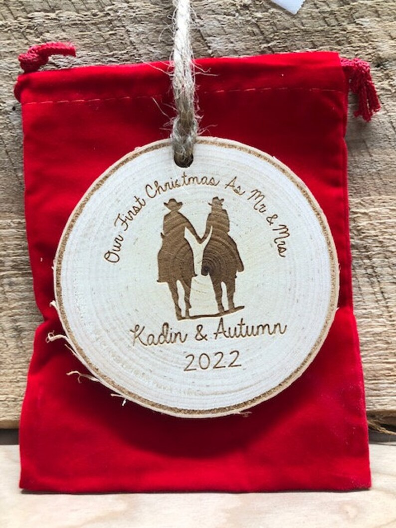 May include: A round wooden ornament with the text "Our First Christmas As Mr & Mrs Kadin & Autumn 2022" and a silhouette of a couple in cowboy hats.