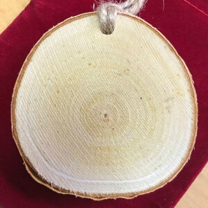 May include: A round wooden ornament with a natural wood grain pattern and a hole in the top for hanging. The ornament is made from a slice of wood and has a rustic, natural look.