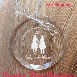 May include: Clear glass Christmas ornament with the text "Our First Christmas as Mr and Mrs 2017 Cody and Allison". The ornament features a silhouette of a couple in cowboy hats riding horses.