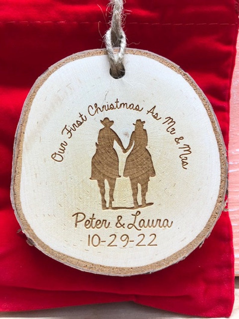 May include: A wooden Christmas ornament with a couple riding horses. The ornament is engraved with the text "Our First Christmas As Mr. & Mrs. Peter & Laura 10-29-22".