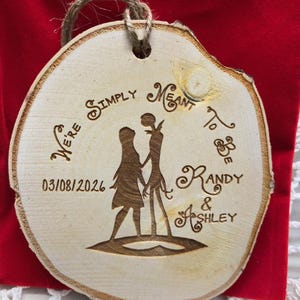 May include: A round wooden ornament with a rustic edge and a jute string for hanging. The ornament features a laser-etched design of a couple holding hands, with the text "We're Simply Meant to Be" and the names "Randy & Ashley" along with the date 03/08/2026.