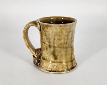 May include: A handmade ceramic mug with a unique, textured surface. The mug features a wide, sturdy handle and a slightly flared shape. The color is a blend of tan and brown, with darker streaks adding visual interest. A rustic, artisanal piece.