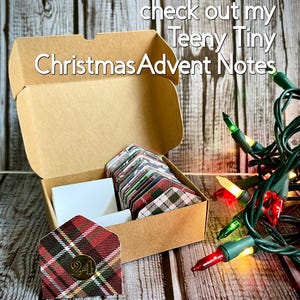 May include: A brown cardboard box with a plaid patterned envelope inside. The box is open and the envelope is visible. The envelope has the number 24 on it. The box is sitting on a wooden surface. There are Christmas lights in the background. The text on the box reads "Visit my shop to check out my Teeny Tiny Christmas Advent Notes".