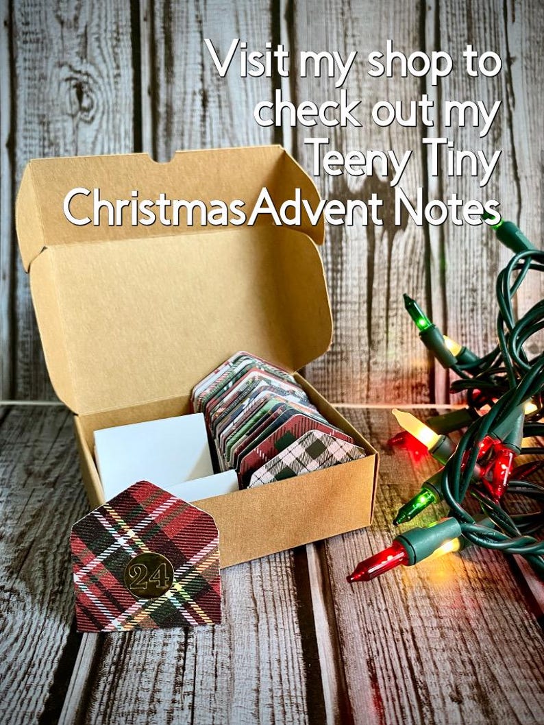 May include: A brown cardboard box with a plaid patterned envelope inside. The box is open and the envelope is visible. The envelope has the number 24 on it. The box is sitting on a wooden surface. There are Christmas lights in the background. The text on the box reads "Visit my shop to check out my Teeny Tiny Christmas Advent Notes".