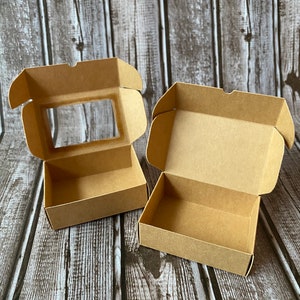 May include: Two open cardboard boxes, one with a clear window, on a woodgrain background. Both boxes are brown and have a simple design.