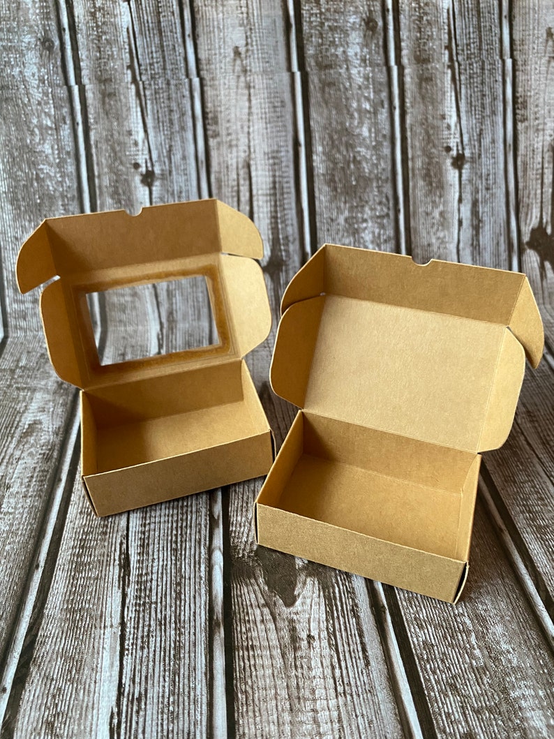 May include: Two open cardboard boxes, one with a clear window, on a woodgrain background. Both boxes are brown and have a simple design.