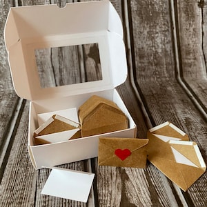 May include: A white box with a clear window, containing a stack of brown envelopes with white lining.  There are several envelopes outside the box, one with a red heart sticker.
