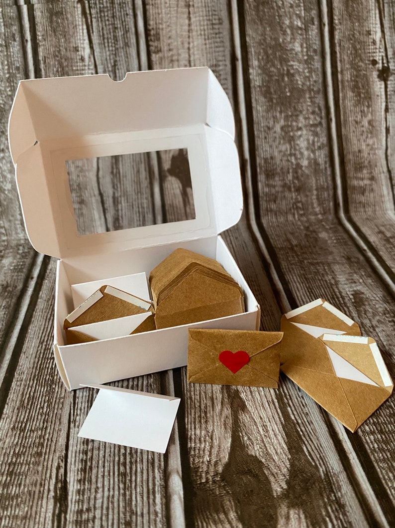 May include: A white box with a clear window, containing a stack of brown envelopes with white lining.  There are several envelopes outside the box, one with a red heart sticker.