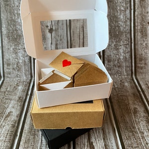 May include: A set of small brown envelopes with red hearts on the front, stacked inside a white box with a clear window. The box is sitting on top of a brown box and a black box.